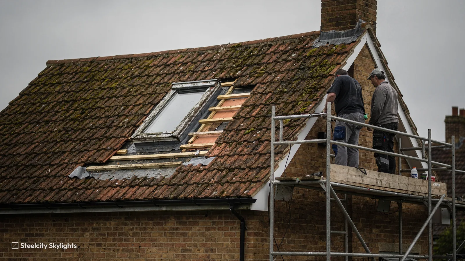 Two roofers on a scaffold beside an old roof window being taken out of a clay tile roof
