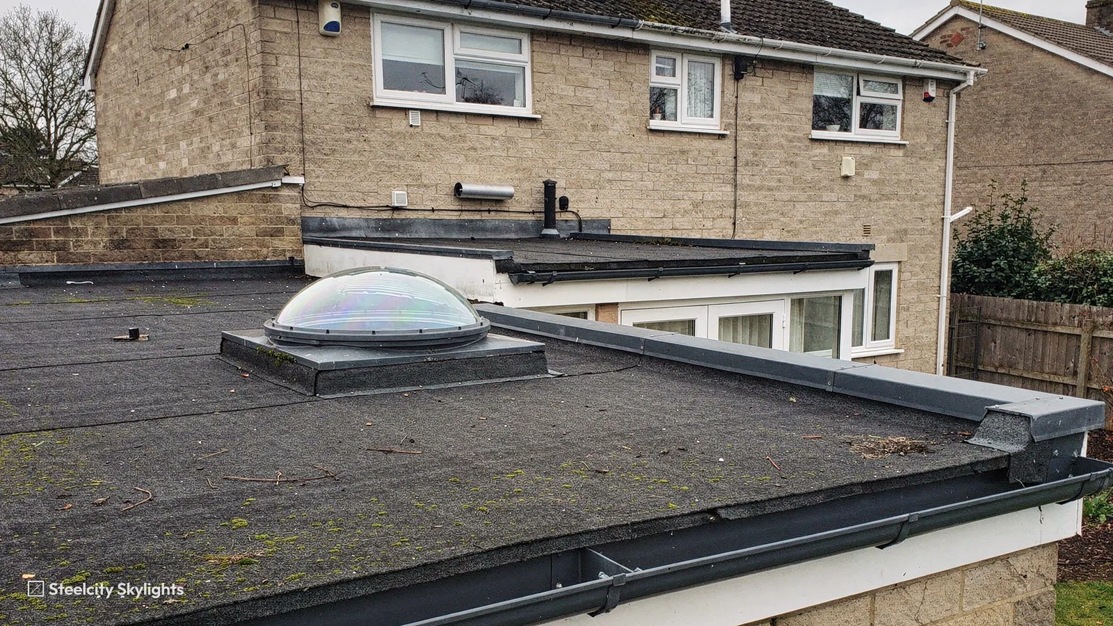 Clear dome of a sun tunnel on a felt flat roof beside a pitched-roof stone house