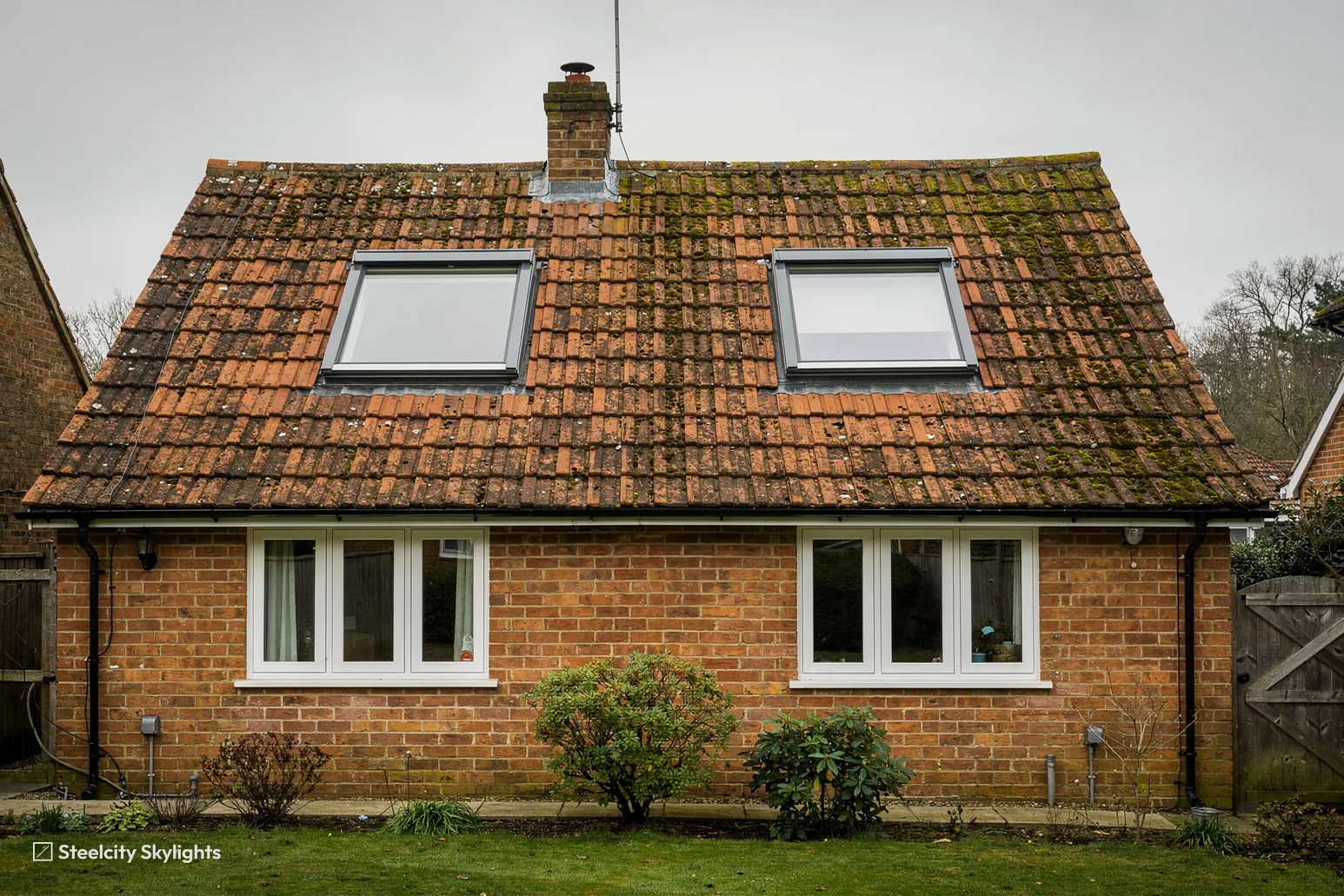Brick cottage with two roof windows in a mossy clay tile roof and a front lawn