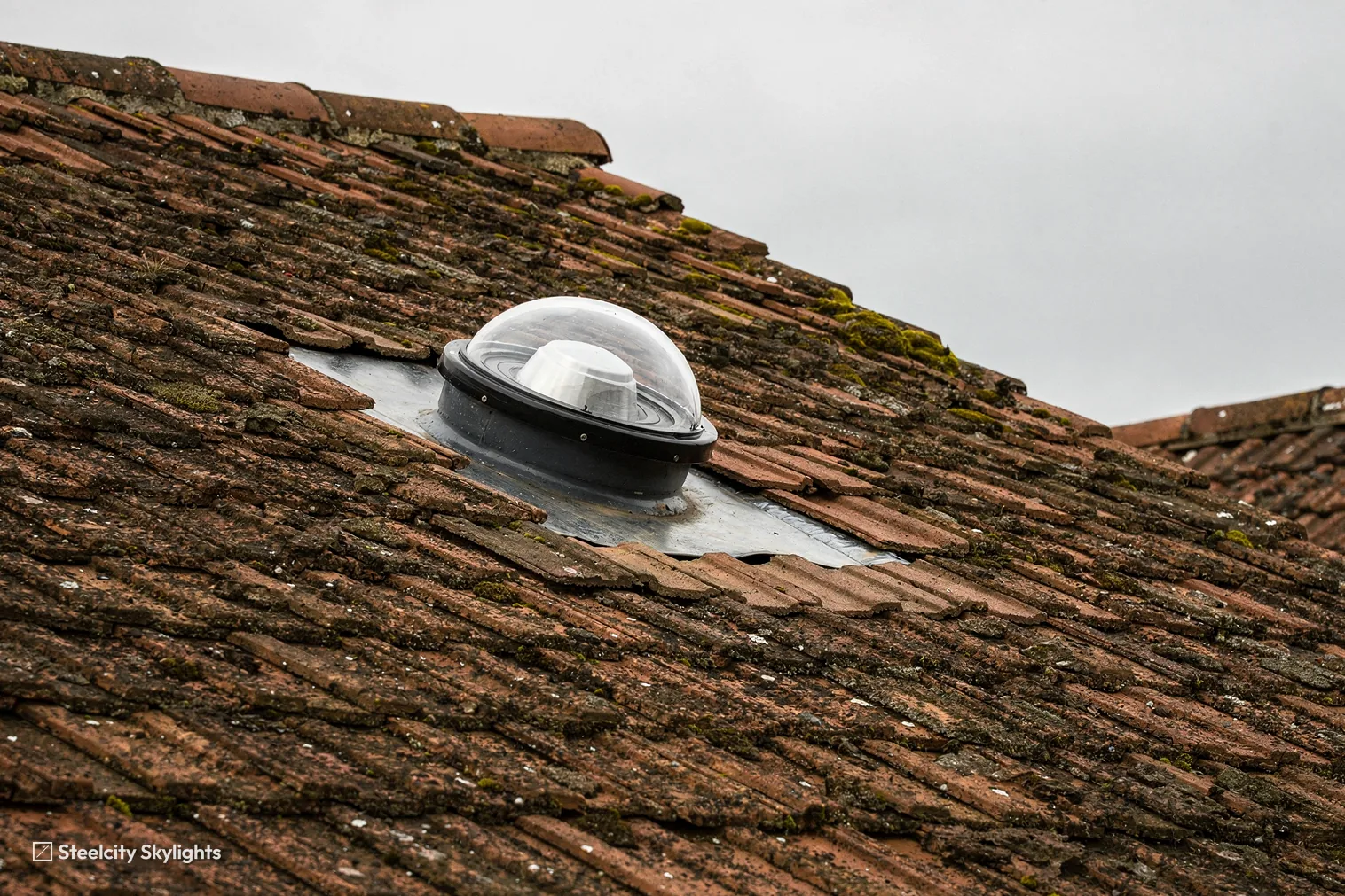 Clear domed sun tunnel cap fitted into a mossy clay tile roof