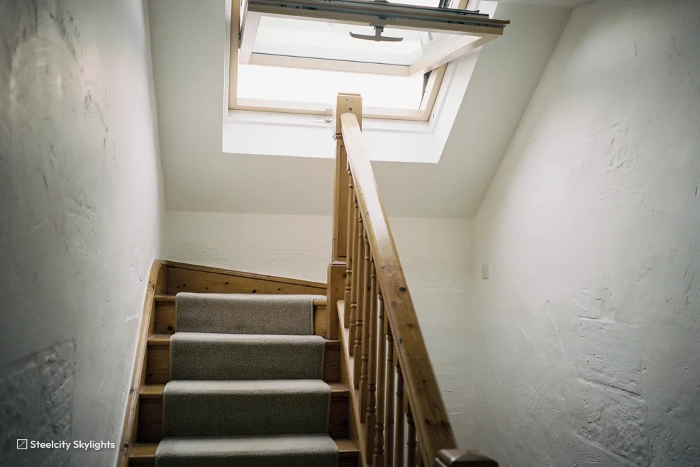 Narrow stairwell with a wooden handrail lit by an open roof window at the top