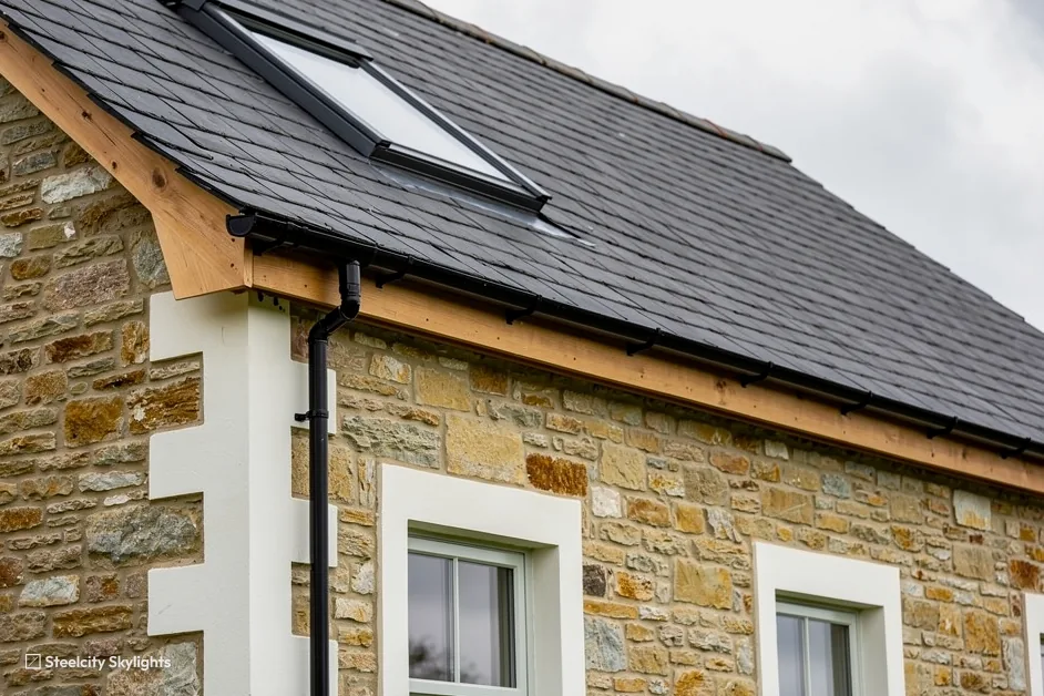 Roof window in a dark slate roof above a stone-built house with a timber fascia