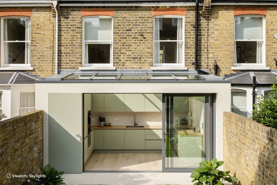 Rear of a yellow brick terrace with a glass-roofed extension open onto a sage green kitchen
